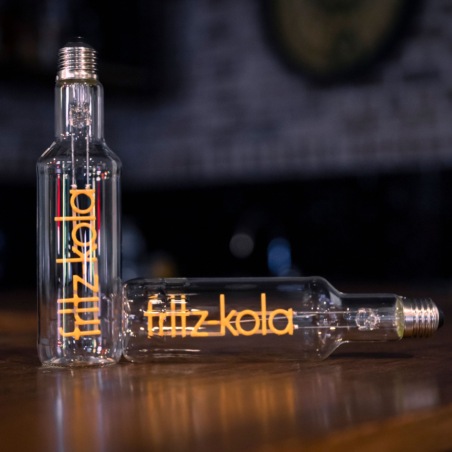 Zwei Glühbirnen mit orangefarbenem fritz-kola Schriftzug als Glühfaden  auf Holztisch, eine aufrecht, eine liegend, vor dunklem Hintergrund mit unscharfen Lichtern.