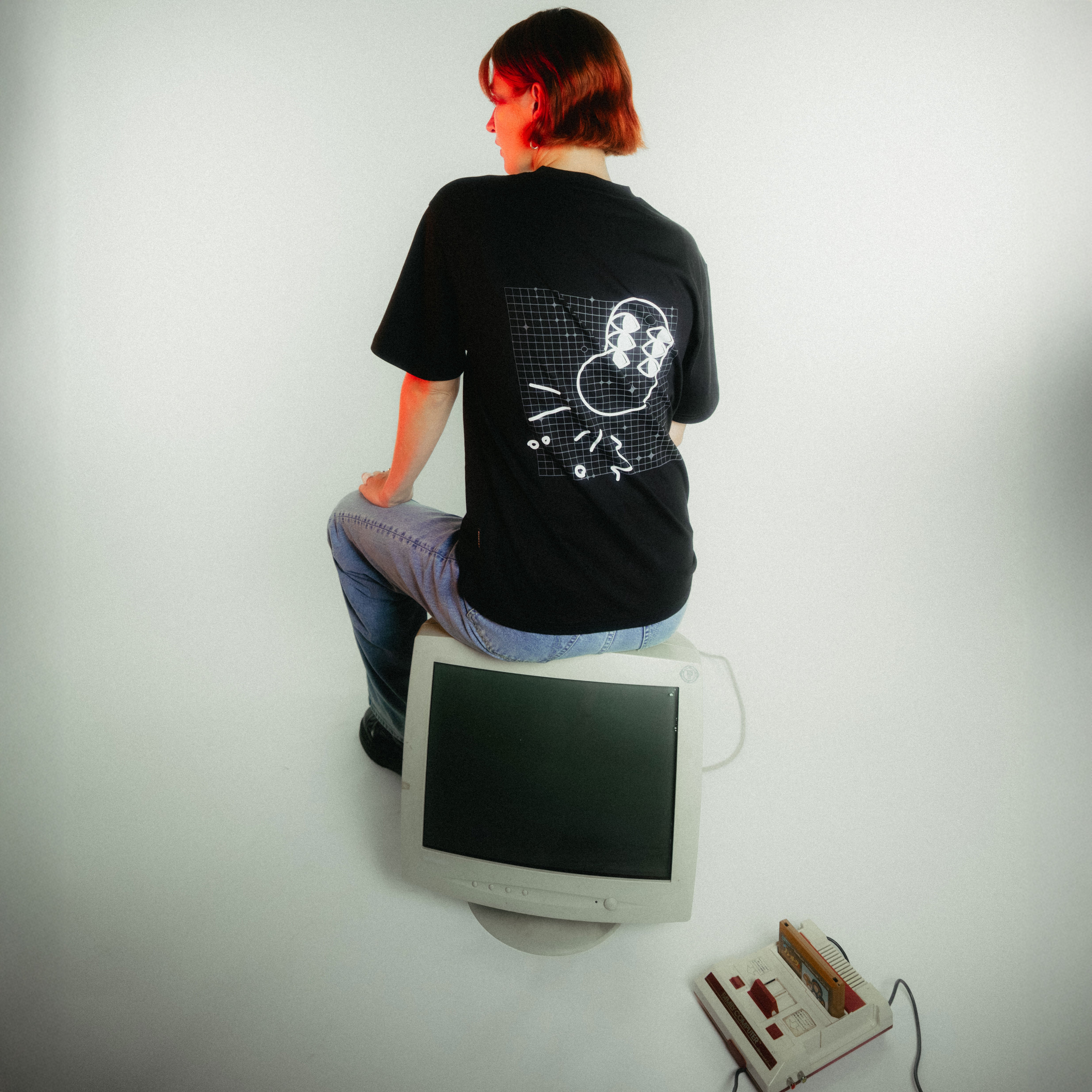 Frau mit kupferfarbenem Bob sitzt von hinten auf weißem Retro-Monitor, trägt schwarzes T-Shirt mit von fritz-kola x rktbns - weiße Bohne mit 6 Augen  und Gitterlinien auf schwarzem Grund.