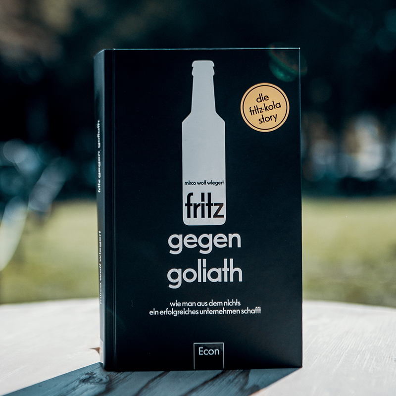 Schwarzes Buch "fritz gegen goliath" von mirco wiegert steht aufrecht. Cover zeigt weiße Flaschensilhouette mit "fritz" Schriftzug. Goldener Aufkleber "die fritzkola story" oben rechts. Untertitel "wie man aus dem nichts ein erfolgreiches unternehmen schafft". Econ Verlag Logo unten.