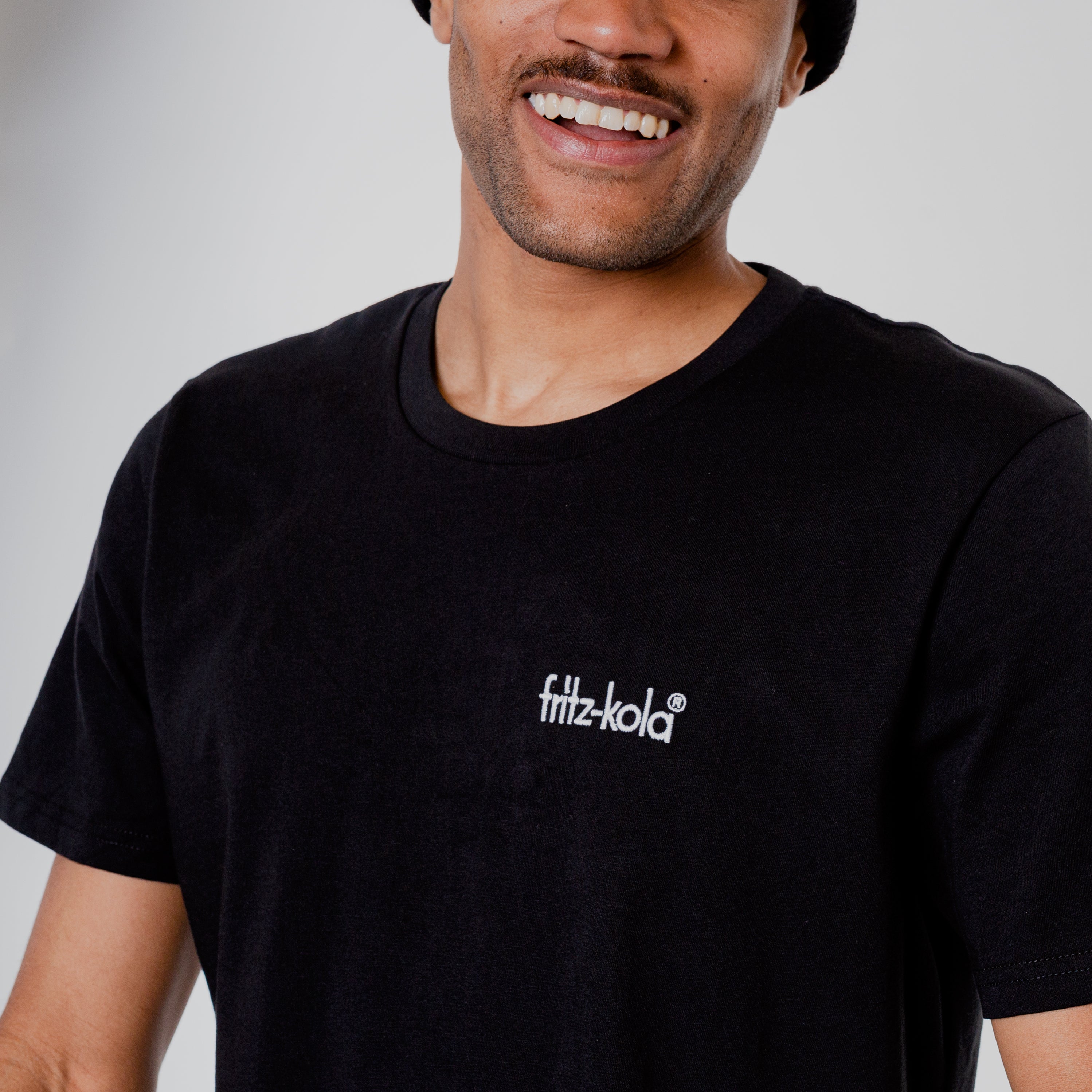 Nahaufnahme eines lächelnden Mannes in schwarzem T-Shirt mit kleinem weißem "fritz-kola" Schriftzug auf der Brust. 