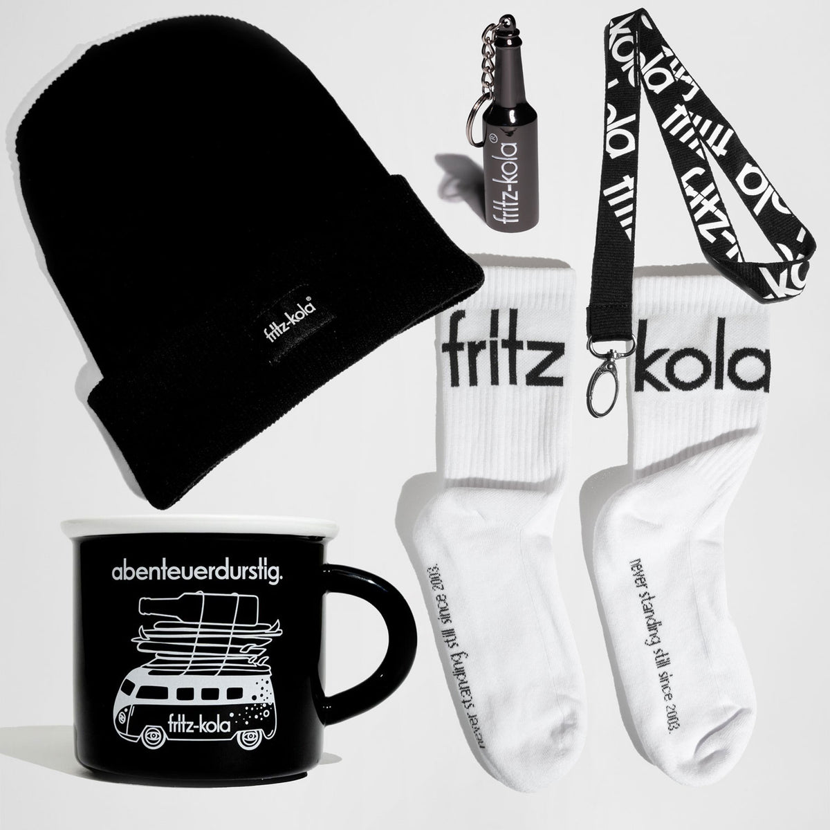 fritz-kola Geschenkset „fritzfest“ auf weißem Hintergrund mit schwarzer Strickmütze mit kleinem fritz-kola Logo, weißen Socken mit großem schwarzem fritz-kola Schriftzug, schwarzer Tasse mit weißer „abenteuerdurstig“-Aufschrift und Busmotiv sowie schwarzem Schlüsselband und kleinem Flaschen-Schlüsselanhänger im fritz-kola Design.