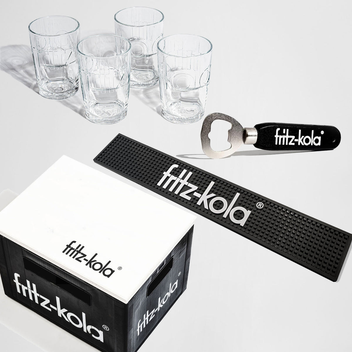 Geschenkset von fritz-kola auf weißem Hintergrund. Zu sehen sind vier transparente Trinkgläser mit eingeprägtem fritz-kola Logo, ein Flaschenöffner mit schwarzem Griff und weißem fritz-kola Schriftzug, eine längliche schwarze Barmatte mit großem weißem fritz-kola Logo sowie eine schwarze Getränkekiste mit einer weißen Sitzauflage aus Holz.
