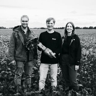 Auf einer schwarz-weiß Fotografie stehen drei Personen, darunter fritz-kola Grüner Mirco Wolf Wiegert auf einem Feld. Mirco hat dabei eine große Zuckerrübe in der Hand und zeigt sie lächelnd in die Kamera